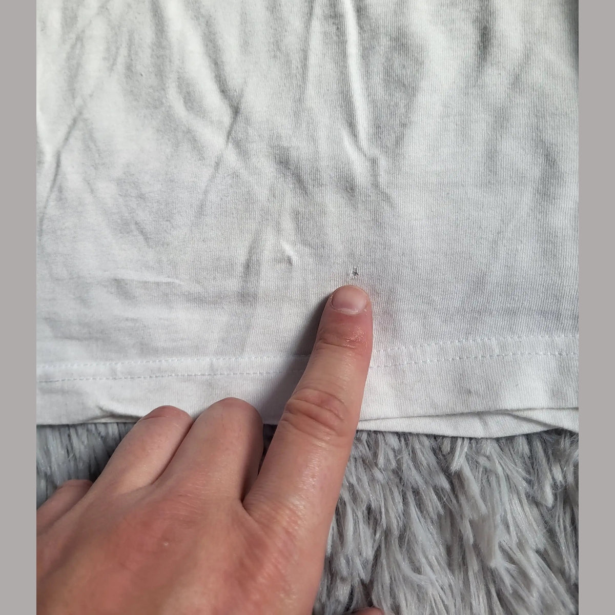 T-shirt blanc, marque et taille non visibles, petit trou montré du doigt. Fripes Sélect