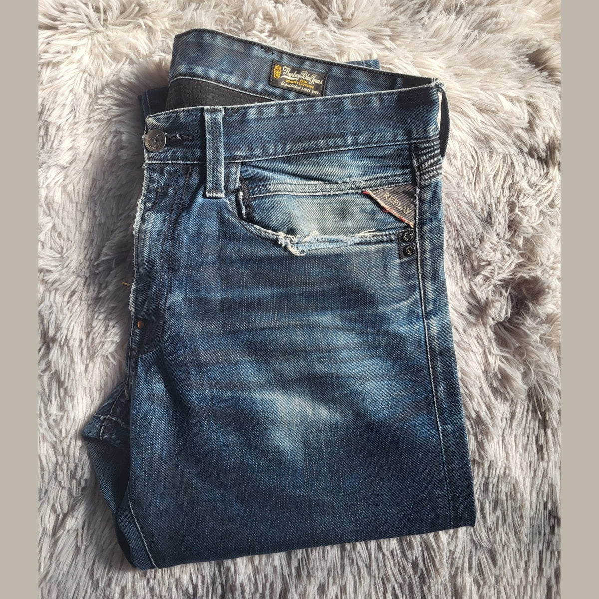 Jean bleu taille 32 de la marque Replay, plié sur un tapis duveteux. Fripes Sélect